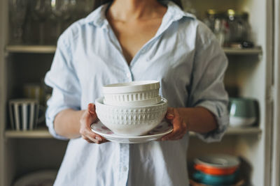 Young smiling asian woman in casual shirt with dishes near vintage sideboard at home