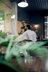 Side view full body of bearded barber spraying hair of male customer sitting on chair in modern barbershop during procedure