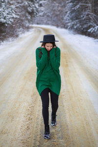 A young girl in a green sweater stands in the middle of a snowy road in a thick pine forest. f