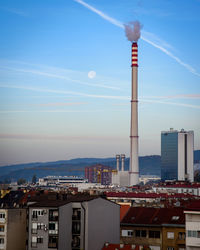 A view of the blue sky with contrails, moon and a heating plant chimney above a city skyline