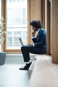 Young businessman using laptop while sitting against window at workplace