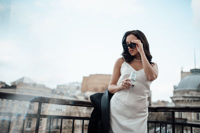 Low angle view of woman holding sunglasses and champagne flute standing by railing against sky