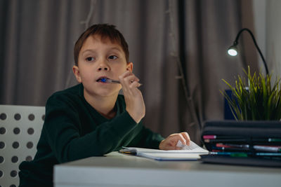 Boy biting pen looking away sitting at desk