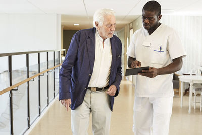 Male nurse using digital tablet with senior male patient at hospital corridor