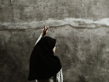 Rear view of woman gesturing peace sign while standing against wall