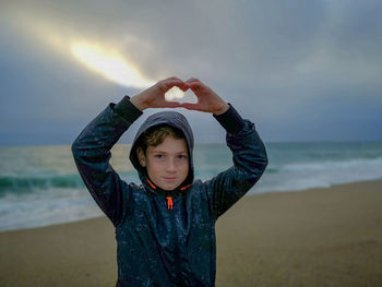 Portrait of boy with arms raised making heart shape at beach against sky