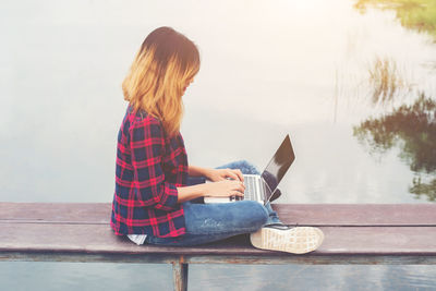 Side view of woman using laptop while sitting on retaining wall