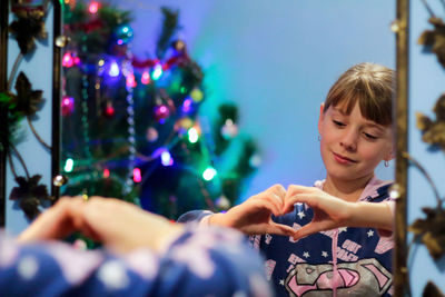 Little blonde caucasian girl 10 years old in pajamas shows gesture heart on mirror reflection.
