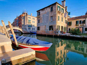 Boats moored in canal by buildings against blue sky