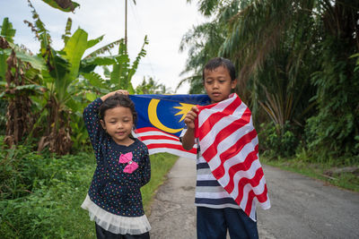 Siblings holding malaysian flag while standing on road amidst plants