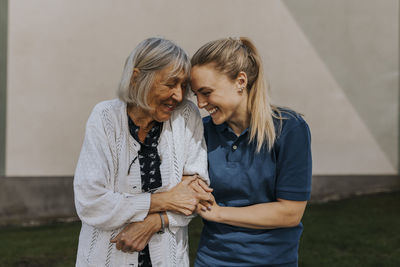 Happy female caregiver with arm in arm of senior woman standing against wall