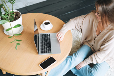 Young smiling woman in beige cardigan and jeans using laptop in room, top view