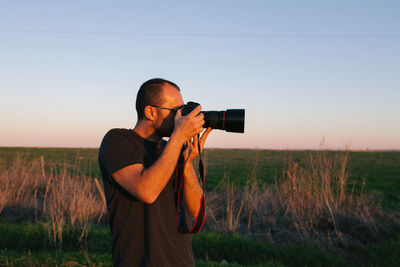 Man holding camera while standing on field against sky during sunset