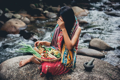 Smiling woman holding fresh food in bucket while sitting on rock amidst river