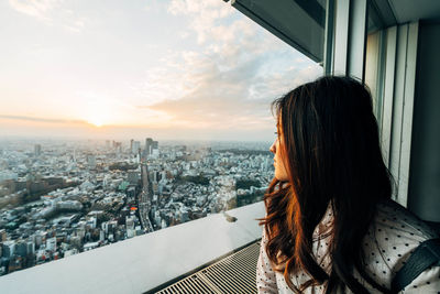 Woman looking at city through window against sky