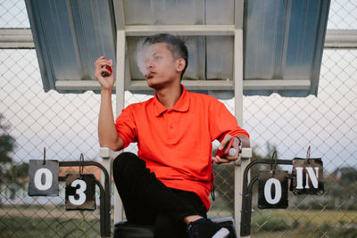 Young man smoking electronic cigarette while sitting on tennis court