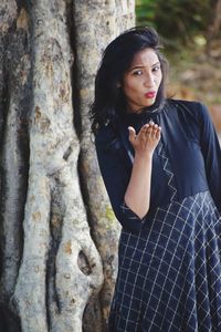 Portrait of woman blowing kiss while standing against tree trunk