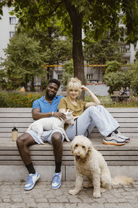 Portrait of smiling man and woman sitting with dogs on bench in public park