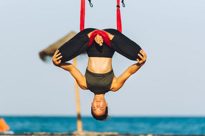 Woman practicing aerial yoga on silk at beach against sky