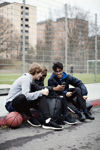 Full length of social media addicted friends wearing warm clothing while sitting on sidewalk after basketball practice d