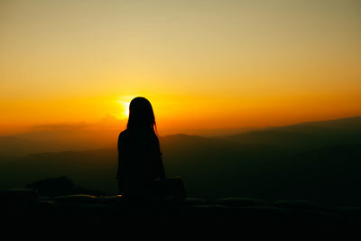 Silhouette woman sitting against orange sky