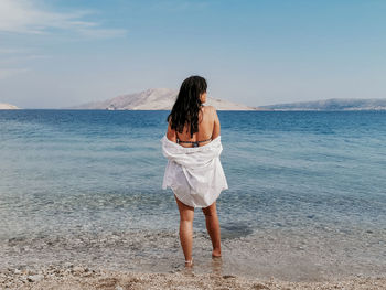 Rear view lifestyle image of stylish young woman in white shirt standing on beach and looking at sea