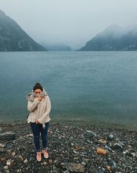 Full length of woman standing on stones against lake and mountains during winter