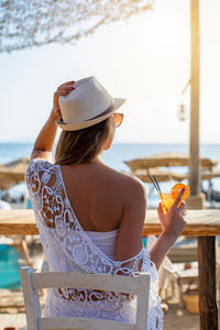 Rear view of young woman holding drink while sitting on cafe at beach against clear sky