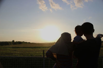 Rear view of family standing on field against sky during sunset