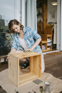 Young woman making wooden furniture while kneeling on porch outside house