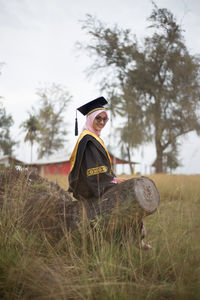 Portrait of woman in graduation gown sitting on log