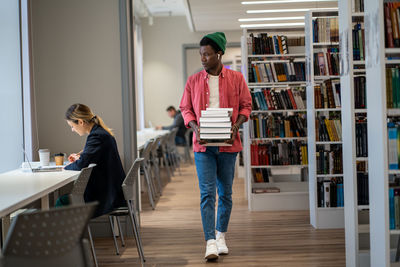 African american international student guy walking with stacked books through college library