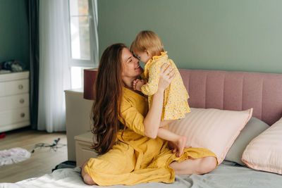 Young red-haired mother plays with her little cheerful daughter sitting on the bed.