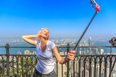 Cheerful woman taking selfie while standing against clear sky in balcony