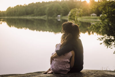 Mother with arm around daughter sitting on rock near lake