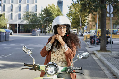 Young woman fastening helmet while sitting on motor scooter in city