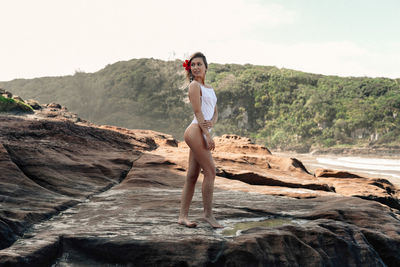 Seductive young woman wearing white swimsuit while standing on rock at beach