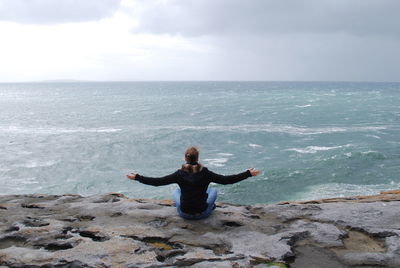 Rear view of woman with arms outstretched sitting on cliff by sea against sky