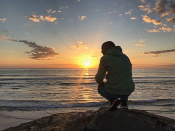 Rear view full length of man crouching on rock at beach during sunset