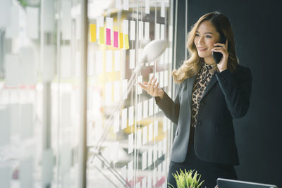 Smiling businesswoman talking over smart phone while standing by window in office