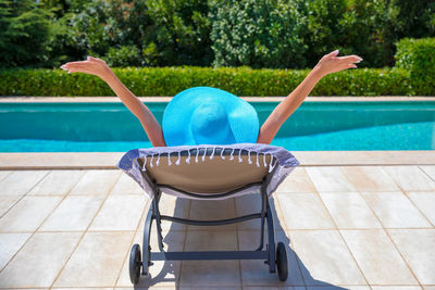 Rear view of woman with arms outstretched relaxing on lounge chair at poolside