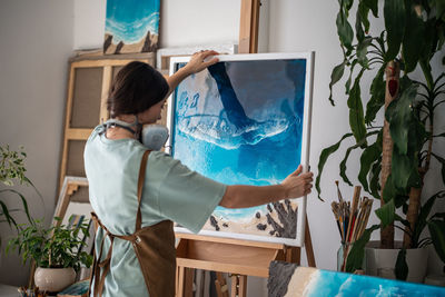 Thoughtful creative woman artist places finished epoxy painting on wooden easel in art workshop.