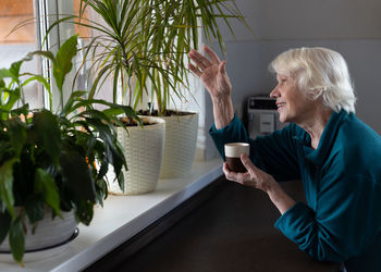 An attractive elderly woman drinks espresso in the kitchen and looks out the window