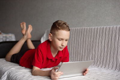 A cute boy in a red t-shirt is lying on the couch and playing with a tablet
