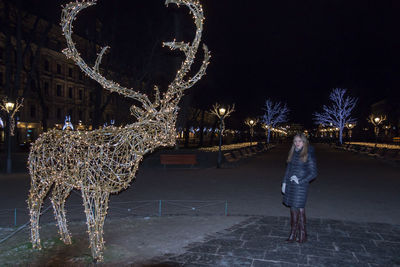 Woman standing by illuminated artificial deer at night