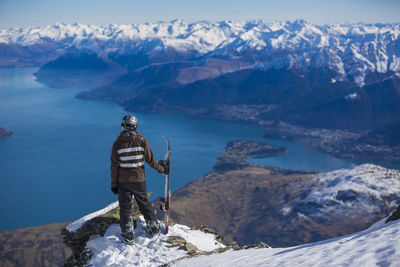 Rear view of person with snowboard standing on cliff by sea against mountains during winter