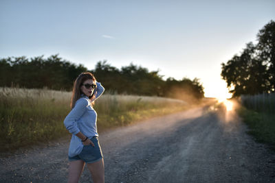 Young woman walking on dirt road by field against sky during sunset