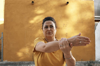 A middle-aged woman is stretching with a yellow wall in the background