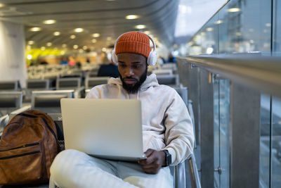 Young hipster black man sitting in airport terminal using laptop wear headphones and listening music