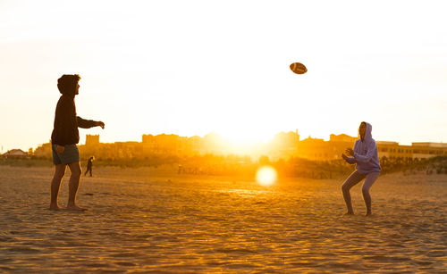 Friends playing with rugby ball at beach against sky during sunset